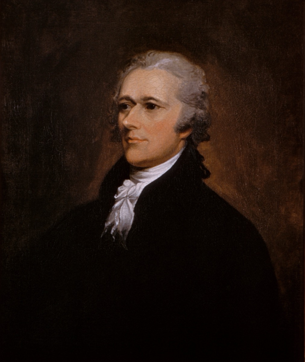 Standing oil portrait of Alexander Hamilton in dark coat and white cravat, hand resting on a desk